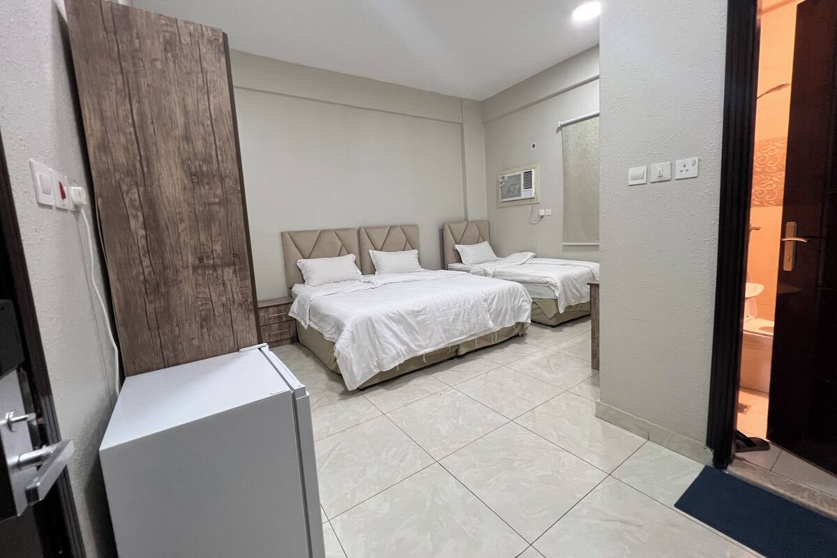 Habitación de hotel con dos camas, un armario de madera, un mueble blanco y un baño visible a través de una puerta abierta.