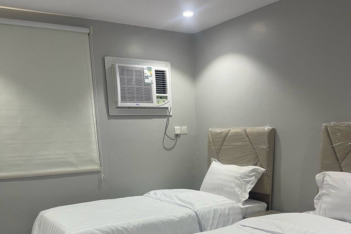 Habitación de hotel con dos camas, una ventana con persianas, un equipo de aire acondicionado y piso de baldosas.
