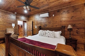Una habitación con paneles de madera, una cama, un ventilador de techo, un aire acondicionado, una silla y una mesita de noche.
