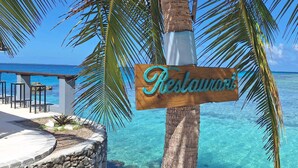 Uma placa de madeira com a palavra 'Restaurant' escrita, cercada por palmeiras e vista para o oceano.