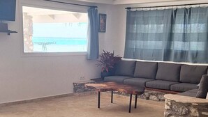 Uma sala de estar com sofá, mesa de centro, tapete estampado, parede de pedra e vista para o mar através de uma janela.