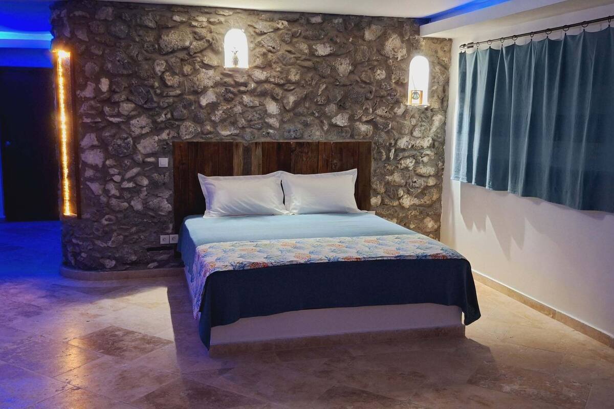 Un dormitorio con pared de piedra, estructura de cama de madera, colcha estampada y cortina azul.