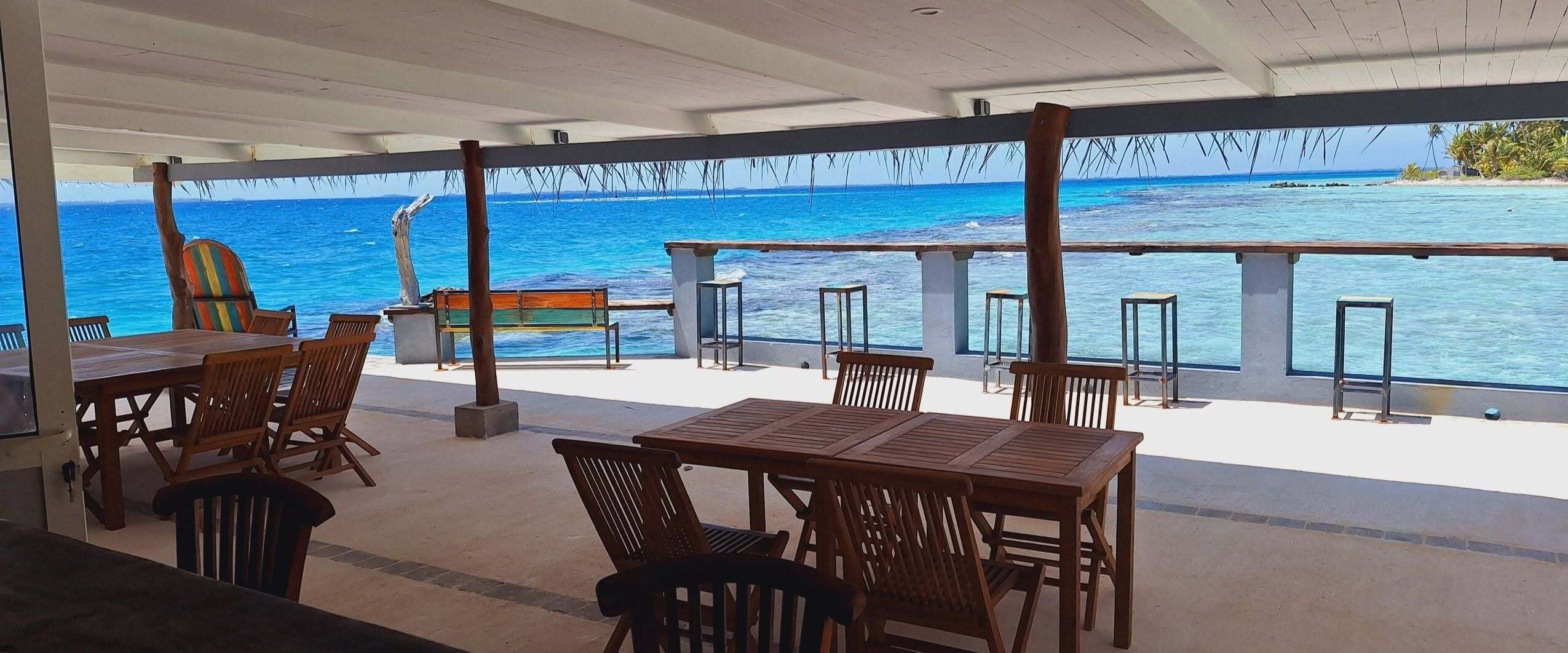 Un'area salotto all'aperto con tavoli e sedie in legno, con vista su un mare cristallino e un'isola tropicale.