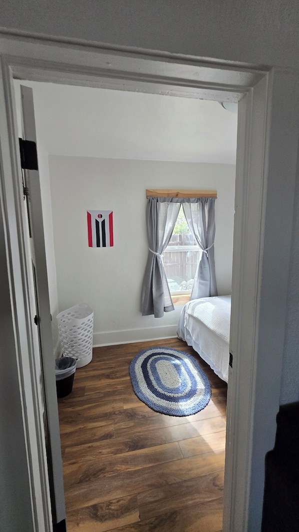 A bedroom with a wooden floor, a bed, a window with curtains, a rug, and a wall-mounted light switch.