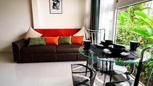 A modern living room with a glass dining table, a brown sofa with colorful cushions, and a view of lush greenery through large windows.