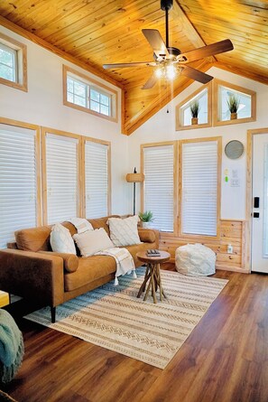 A spacious living room with a wooden ceiling, large windows with blinds, a brown sofa, a small round table, and a decorative rug. - Cozy 1 bedroom 1 bath with loft  tiny home 2 pools & hot tub Lake Travis (Austin)