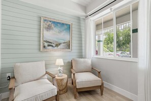 Ein Zimmer mit zwei Stühlen, einem kleinen Tisch mit einer Lampe, einem Bild an der Wand und einem Fenster mit Blick auf Bäume und Autos.