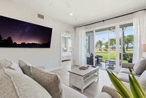 A modern living room with a large window, a flat-screen TV, and a dining area with a table and chairs. - 314 - Renovated First Floor with Golf Views (Naples)