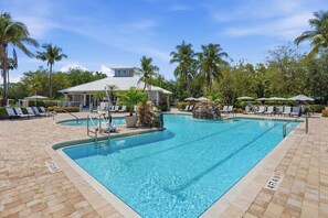 Ein Swimmingpool mit Liegestühlen und Sonnenschirmen, umgeben von Palmen und einem Gebäude mit weißem Dach.