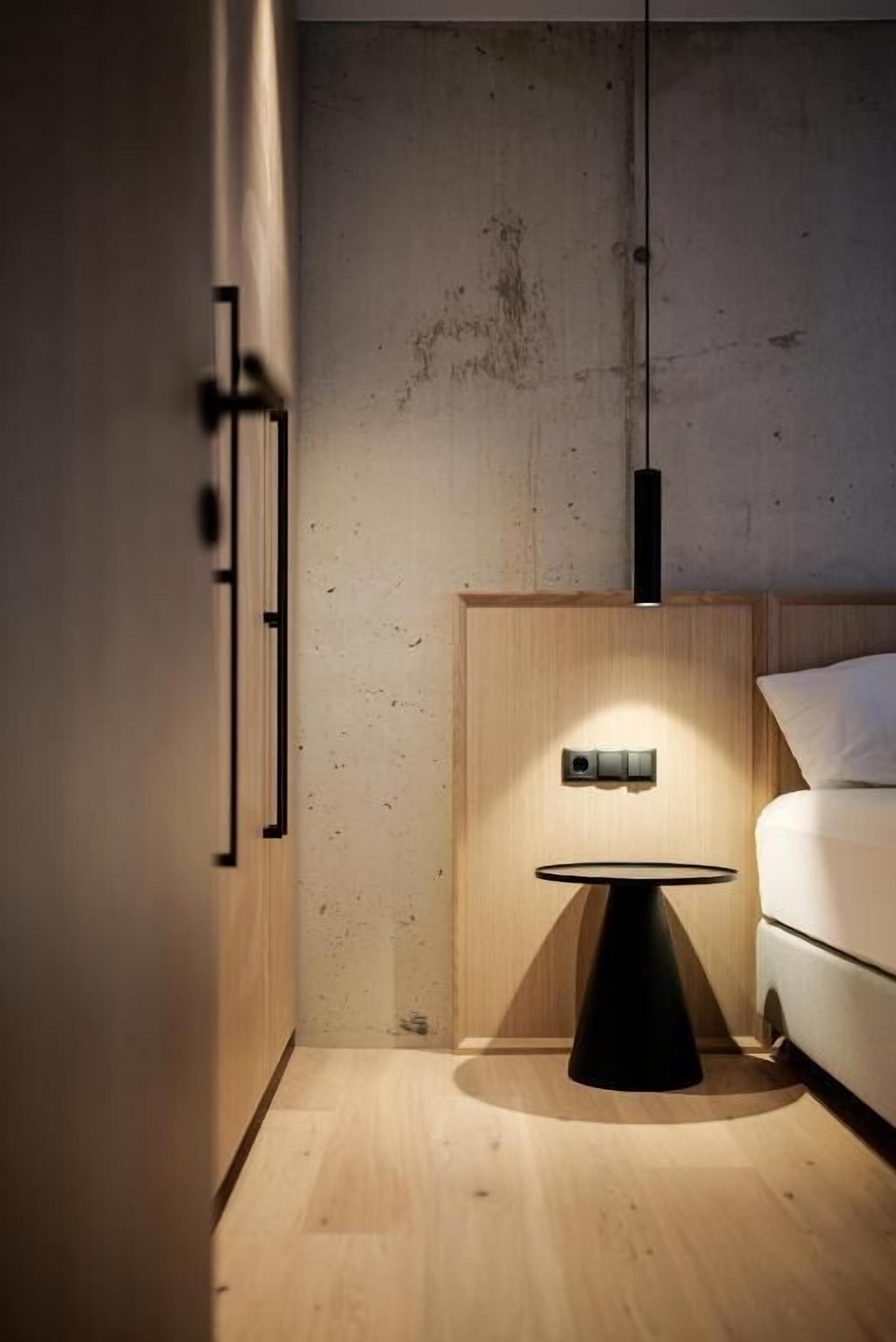Une chambre moderne avec un plancher en bois, une petite table ronde dotée d’un pied noir, un luminaire fixé au mur et un lit avec une literie blanche.