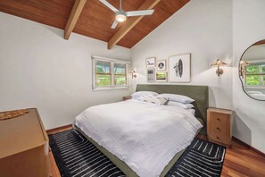 Ein Schlafzimmer mit einem Bett, Deckenventilator, Fenster mit Jalousien, Wandbild, Nachttischlampe und einem Spiegel.