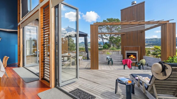 A modern outdoor deck with a fireplace, seating, and a view of the landscape. - Ocean Breeze Family Haven - Pauanui Holiday Home (Pauanui)