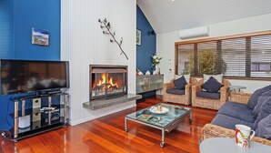 A living room with a fireplace, a flat-screen TV, wicker furniture, and a glass coffee table. - Ocean Breeze Family Haven - Pauanui Holiday Home (Pauanui)