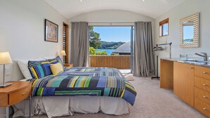 A bedroom with a bed, bedside table, lamp, dresser, and a view of the outdoors. - Ocean Breeze Family Haven - Pauanui Holiday Home (Pauanui)