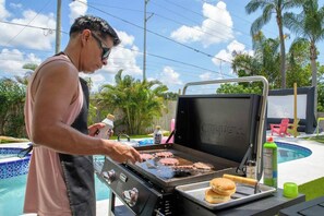 Ein Mann grillt Burger draußen, im Hintergrund ein Pool und Palmen.