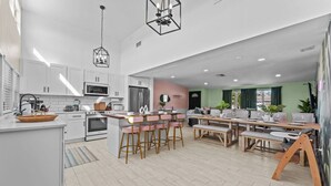 Eine moderne Küche mit weißen Schränken, einer zentralen Kochinsel mit Barhocker, einem Essbereich mit Holztisch und einem Wohnzimmer mit Sofa und grünen Wänden.