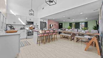 A modern kitchen with white cabinets, a central island with bar stools, a dining area with a wooden table, and a living room with a sofa and green walls.