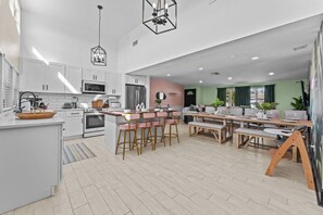 A modern kitchen with white cabinets, a central island with bar stools, a dining area with a wooden table, and a living room with a sofa and green walls. - Heated Pool~Spa~Mini Golf~Fire Pit~Game Room (Clearwater)