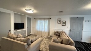 A living room with a flat-screen TV, two beige sofas, a patterned rug, a white door, and a ceiling light. - Quiet Country Retreat East of Nappanee — Relax, Recharge, and Unwind (Nappanee)