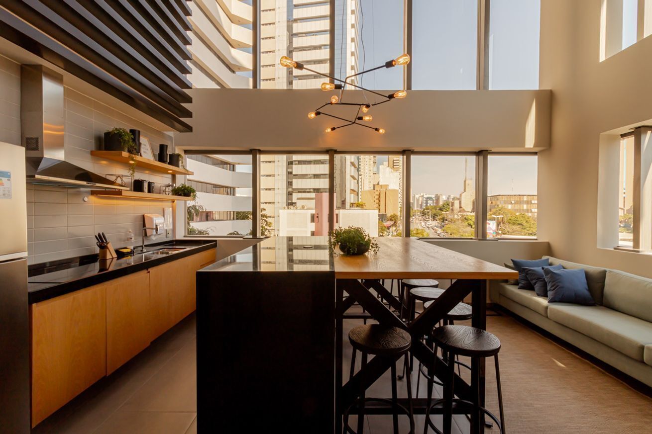 A modern kitchen with a central island, bar stools, a sofa, and large windows offering a city view.