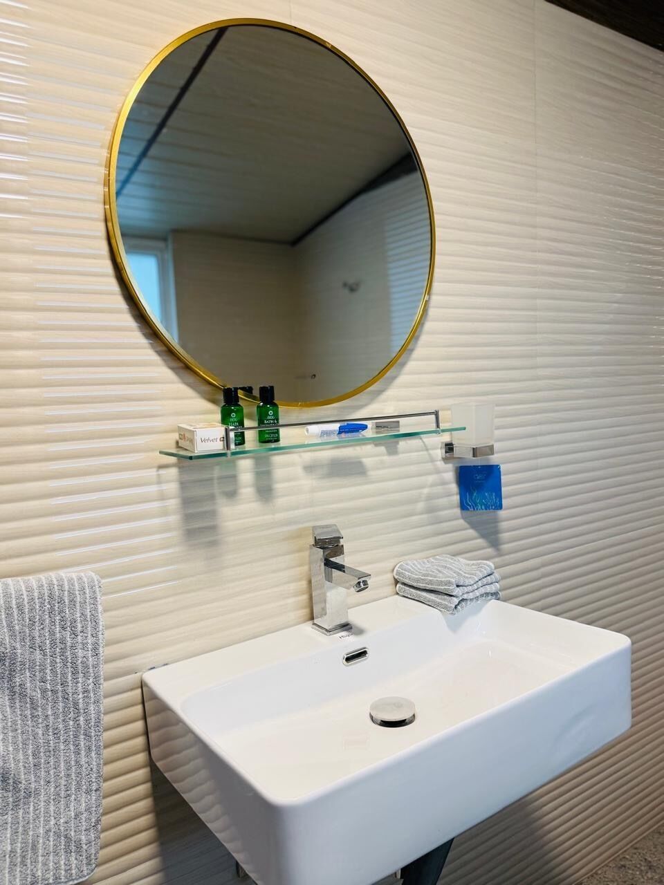 A bathroom with a white sink, a round mirror with a gold frame, and a glass shelf with toiletries.