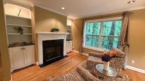 A living room with a fireplace, wooden flooring, patterned furniture, and a window with a view of trees. - NEW 5BR 10-12 guests Fenced Yard, BBQ, Fire Pit (Marietta)