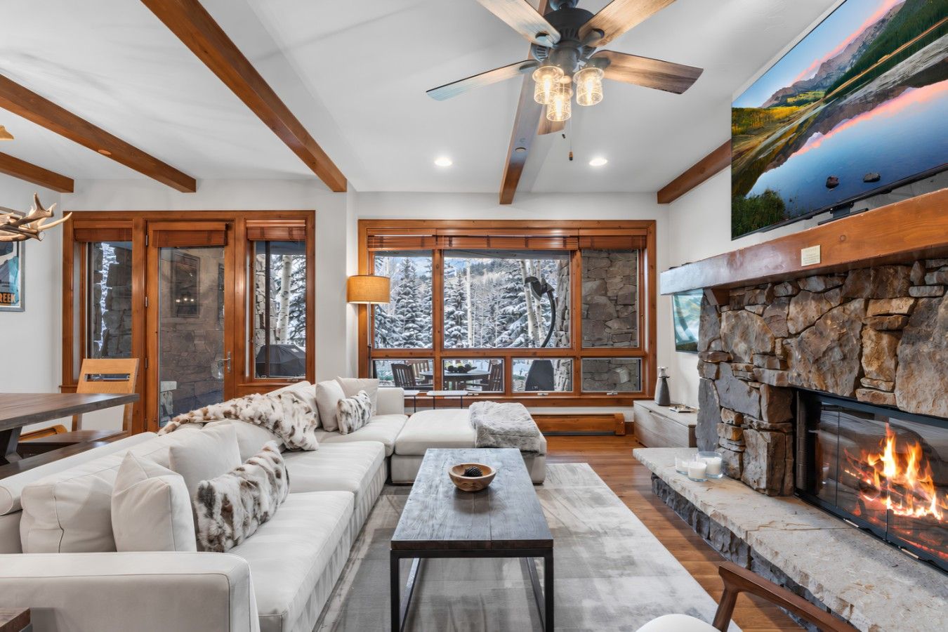 A cozy living room with a fireplace, large windows, and a ceiling fan.
