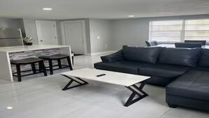 Un salon moderne avec un canapé d’angle, une table basse blanche et un coin cuisine comprenant un bar et des tabourets.