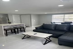 A modern living room with a sectional sofa, a white coffee table, and a kitchen area with a bar and stools. - Cozy 2-bedroom house in perfect Port Richey with AC (Port Richey)
