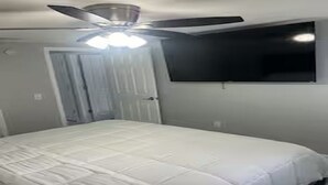 Une chambre à coucher avec un lit, un ventilateur de plafond, un téléviseur fixé au mur et une porte.