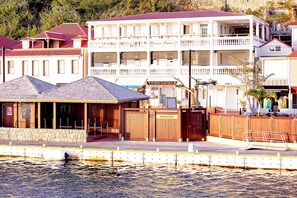 Un hôtel situé au bord de l’eau, avec une façade en pierre, un espace extérieur couvert aménagé en salon et un quai entouré de clôtures.