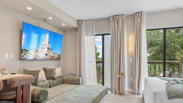 A modern hotel room with a sofa, a bed, a TV, and a balcony with a view of greenery. - Lake View Studio near Universal & Epic (Orlando)