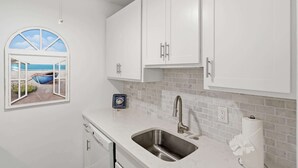 A kitchen with white cabinets, a stainless steel sink, a window with a view of the beach, and a small decorative plate on the counter. - Beach Proximity Condo w/ Pool & Dining Access (Naples)