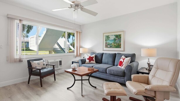 A living room with a sofa, armchairs, a coffee table, a ceiling fan, and a window with a view of buildings and palm trees. - Beach Proximity Condo w/ Pool & Dining Access (Naples)