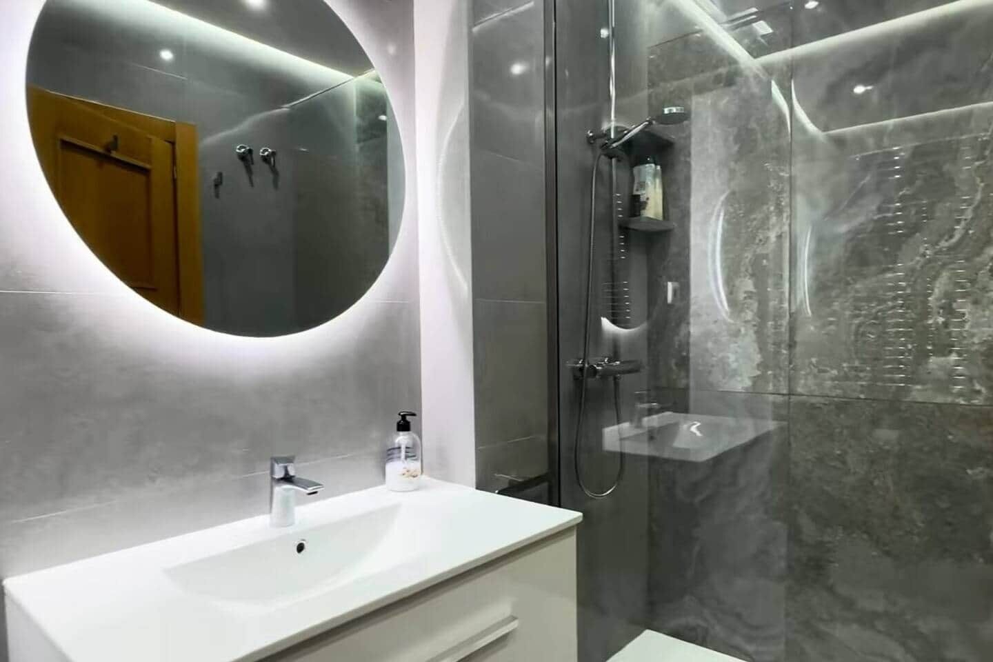 Un baño moderno con un espejo redondo, un lavabo blanco y una ducha con cerramiento de vidrio.