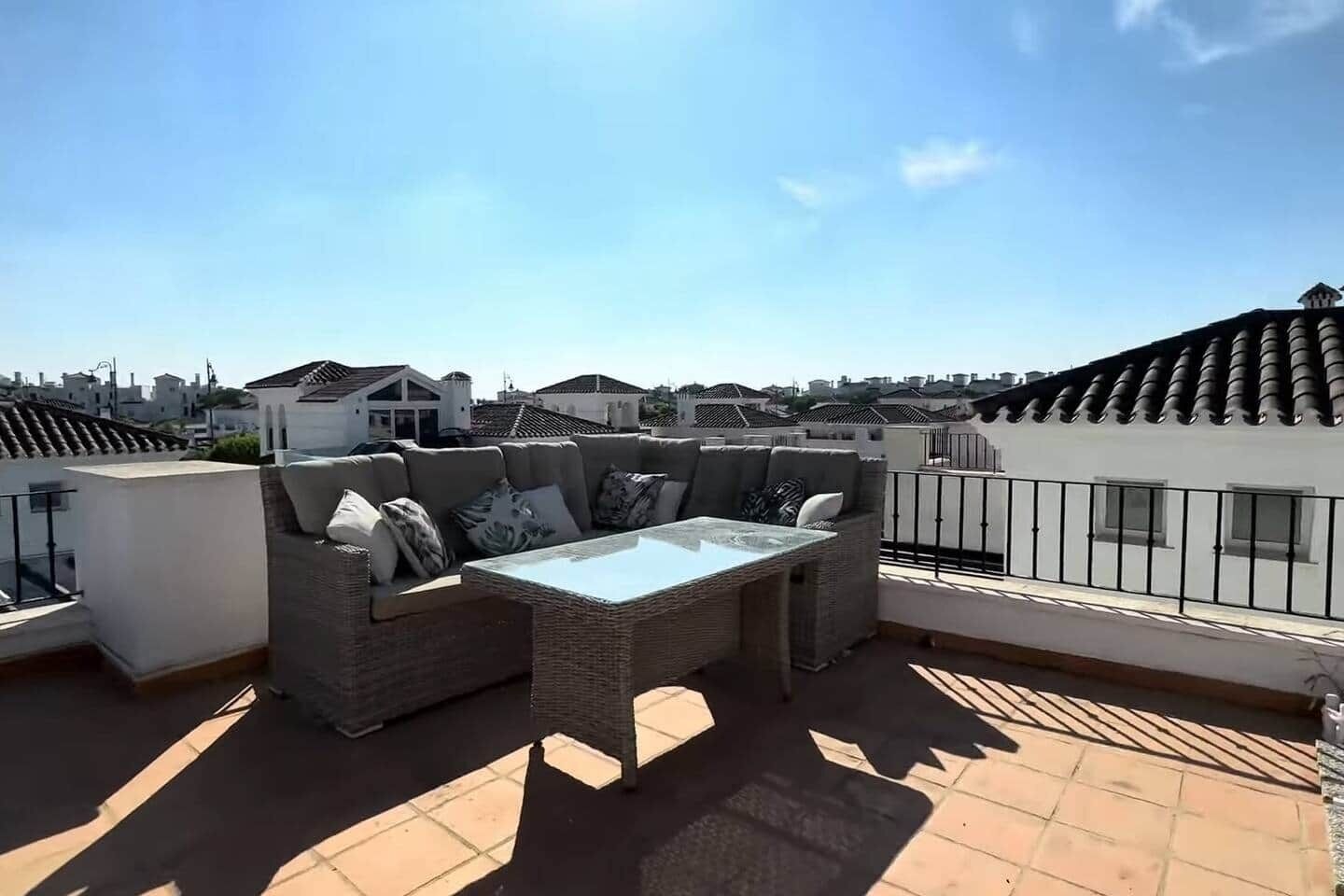 Una terraza en la azotea con un conjunto de sofás de mimbre, una mesa con superficie de vidrio y suelo de baldosas.