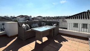 Una terraza en la azotea con un conjunto de sofás de mimbre, una mesa con superficie de vidrio y suelo de baldosas.