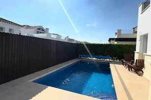 Ein Schwimmbecken mit dunkler Umzäunung, einer Hecke und einigen Liegestühlen.