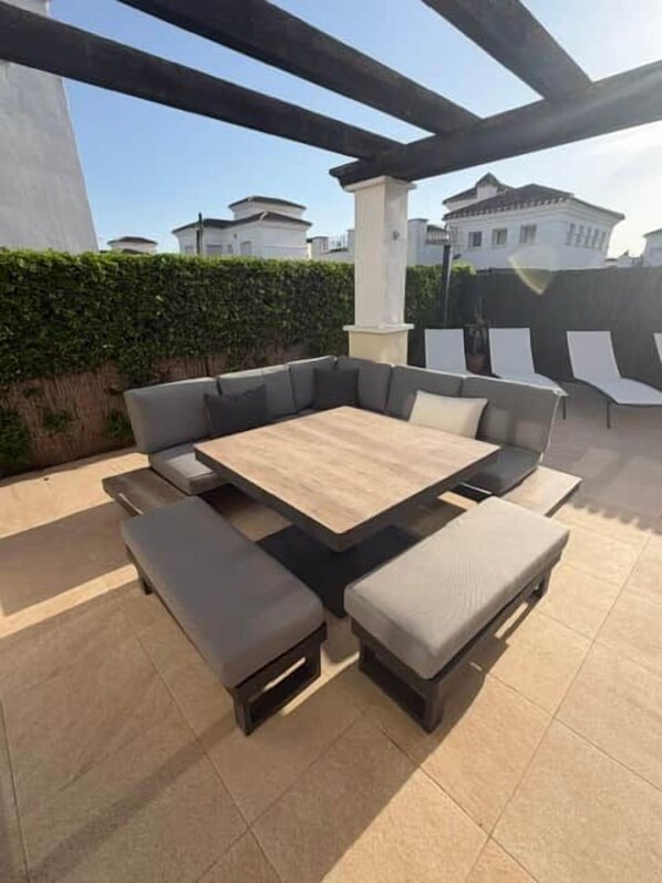 Un patio exterior con una mesa rectangular grande y varias zonas de estar con cojines.