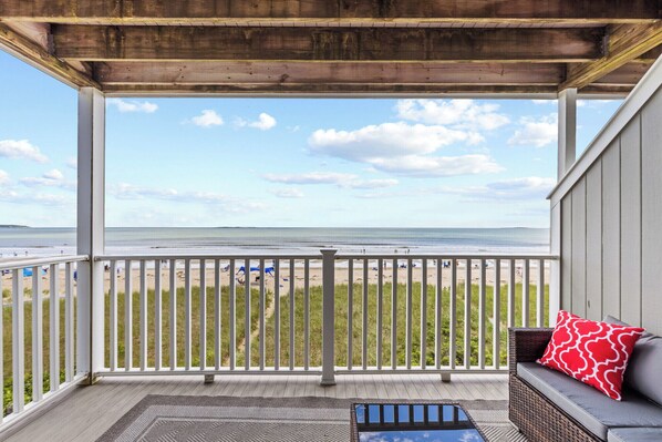 A covered balcony with a view of the beach, a wicker sofa with a red pillow, and a small rug. - 1 Bedroom Beachfront Condo! (Old Orchard Beach)
