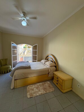 A bedroom with a bed, a nightstand, a chair, a ceiling fan, and a view through an open window. - Cozy house/São Lourenço Pet friendly (São Lourenço)