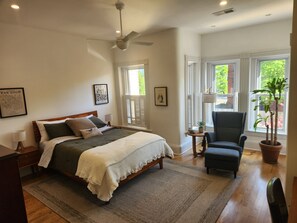 Ein Schlafzimmer mit Bett, Stuhl, Deckenventilator und Fenster mit Blick auf Grünflächen.