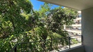 Un balcon doté d’une rambarde en métal, donnant sur une vaste zone verdoyante avec des arbres, et un bâtiment visible au loin.