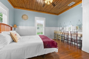 A bedroom with a bed, a wooden ceiling, a dining area with chairs and a table, and a window with blinds.