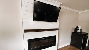 A modern living room with a fireplace, a flat-screen TV mounted above it, and a coffee maker on a black cabinet. - Cozy Resort Condo in Bellaire- Golf, Ski, Lake & Family Fun (Bellaire)