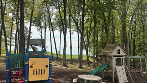A playground with a slide, climbing structures, and a picnic table in a wooded area. - Cozy Resort Condo in Bellaire- Golf, Ski, Lake & Family Fun (Bellaire)
