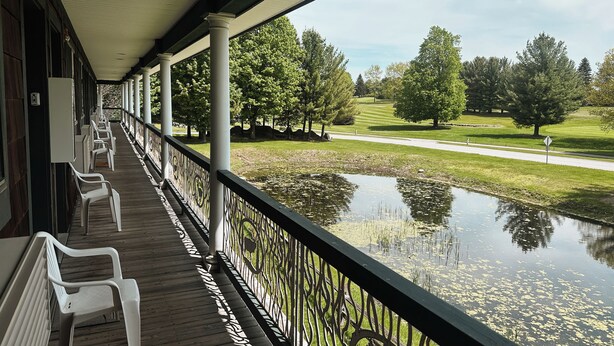 A covered porch with white plastic chairs, a wooden deck, and a view of a pond and golf course. - Cozy Resort Condo in Bellaire- Golf, Ski, Lake & Family Fun (Bellaire)