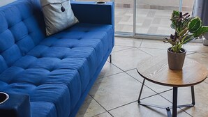 Ein blaues Sofa mit Kissen, ein kleiner Holztisch mit Pflanze und Blick auf eine Stadtlandschaft durch eine Schiebetür.