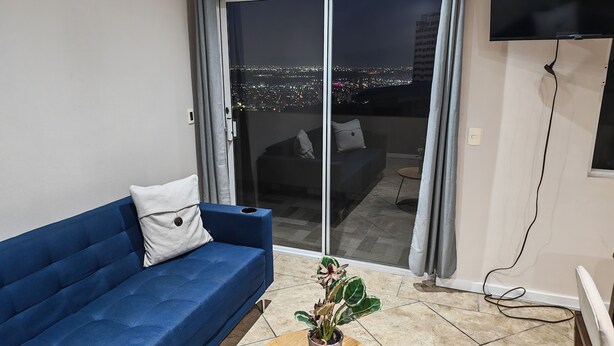 Ein Wohnzimmer mit blauem Sofa, einer Pflanze und einer Glastür, die auf einen Balkon mit Blick auf die Stadt führt.