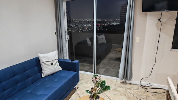 A living room with a blue sofa, a plant, and a sliding glass door leading to a balcony with a city view.